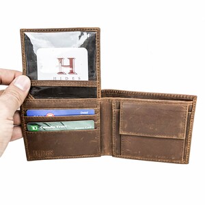 Pu&ograve; includere: Un portafoglio in pelle marrone con una finestra in plastica trasparente per un documento d'identit&agrave;. Il portafoglio &egrave; aperto e ha diverse fessure per carte e un portafoglio per banconote. Il portafoglio ha la parola "HIDES" impressa in rilievo sul davanti.