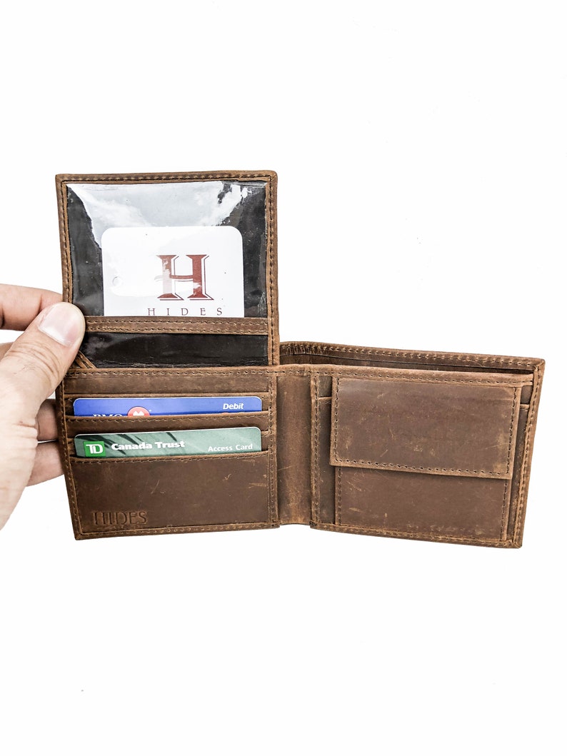 Pu&ograve; includere: Un portafoglio in pelle marrone con una finestra in plastica trasparente per un documento d'identit&agrave;. Il portafoglio &egrave; aperto e ha diverse fessure per carte e un portafoglio per banconote. Il portafoglio ha la parola "HIDES" impressa in rilievo sul davanti.