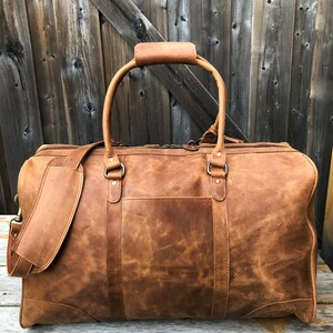 K&ouml;nnte beinhalten: Eine braune Ledertasche mit einem oberen Griff und einem Schultergurt. Die Tasche hat einen Rei&szlig;verschluss und eine Fronttasche.