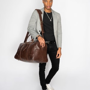 May include: A brown leather duffel bag with a long strap worn over the shoulder of a person wearing a gray blazer, black t-shirt, black jeans, and white sneakers.