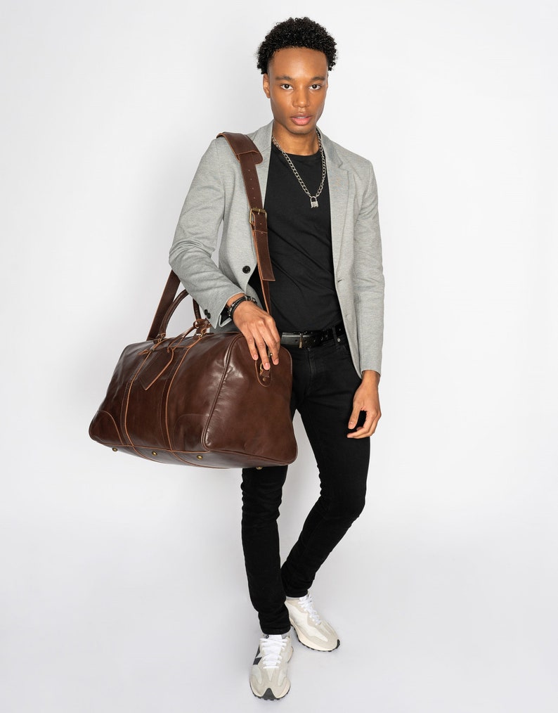 May include: A brown leather duffel bag with a long strap worn over the shoulder of a person wearing a gray blazer, black t-shirt, black jeans, and white sneakers.