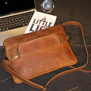 Pode incluir: Uma bolsa de couro marrom para laptop com um bolso com zíper e uma alça de ombro ajustável. A bolsa está sobre uma mesa com um livro e uma esfera de vidro.