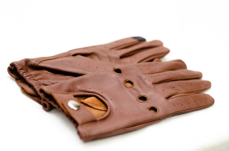 Puede incluir: Un par de guantes de conducir de cuero marr&oacute;n con agujeros en la punta de los dedos y cierre de presi&oacute;n.