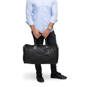 May include: A black woven leather duffel bag with a small zippered pocket on the front. The bag is being held by a person wearing a light blue button-down shirt and dark pants.