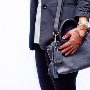 Puede incluir: Un bolso de mano de cuero negro con una correa de cuero marr&oacute;n y un guante de cuero marr&oacute;n. El bolso tiene un borla y una hebilla plateada.
