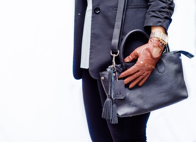 Puede incluir: Un bolso de mano de cuero negro con una correa de cuero marr&oacute;n y un guante de cuero marr&oacute;n. El bolso tiene un borla y una hebilla plateada.