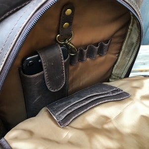 Pu&ograve; includere: Uno zaino in pelle marrone con un'apertura con cerniera, che rivela un interno in tessuto beige con pi&ugrave; tasche. Lo zaino ha un anello per portachiavi e una piccola tasca per un telefono.