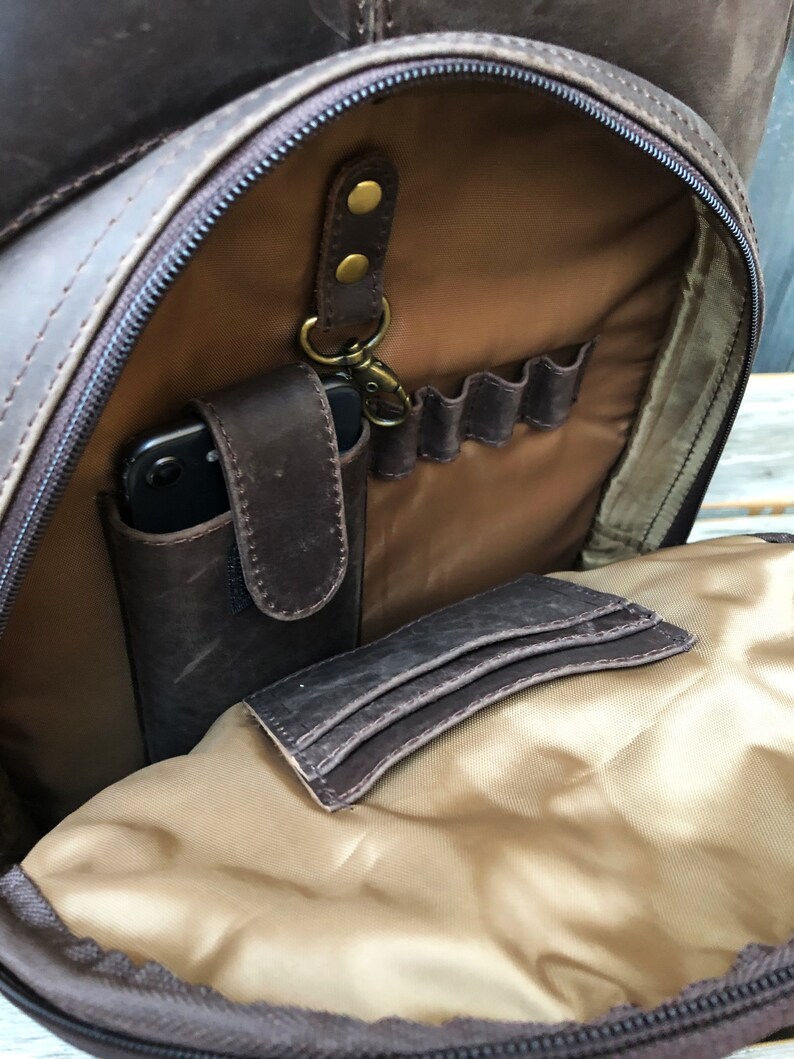 Pu&ograve; includere: Uno zaino in pelle marrone con un'apertura con cerniera, che rivela un interno in tessuto beige con pi&ugrave; tasche. Lo zaino ha un anello per portachiavi e una piccola tasca per un telefono.