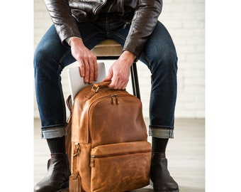 May include: A brown leather backpack with a zippered front pocket and adjustable straps. A person is shown putting a laptop into the backpack.