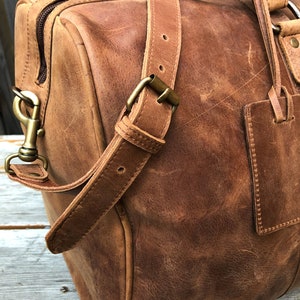 K&ouml;nnte beinhalten: Eine braune Ledertasche mit einem Tragegriff und einem Schultergurt. Die Tasche hat eine Messing-Schnalle und einen Messing-Verschluss.