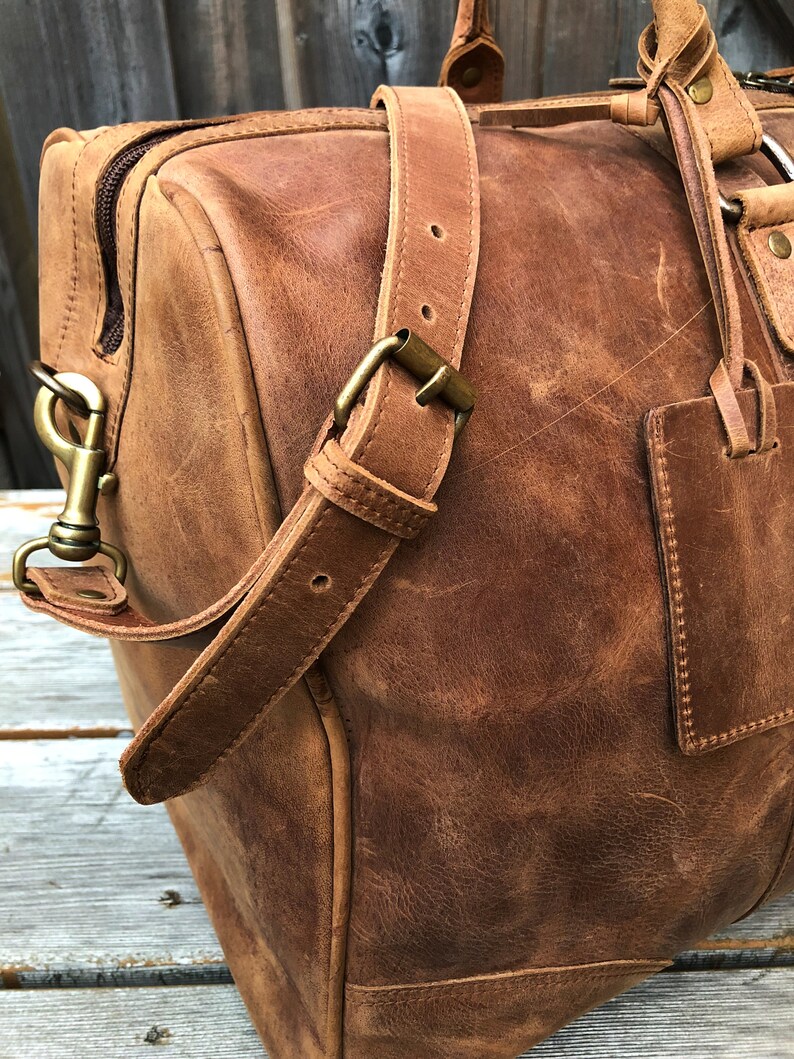 K&ouml;nnte beinhalten: Eine braune Ledertasche mit einem Tragegriff und einem Schultergurt. Die Tasche hat eine Messing-Schnalle und einen Messing-Verschluss.