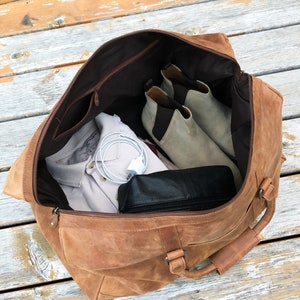 K&ouml;nnte beinhalten: Eine braune Ledertasche mit Rei&szlig;verschluss und einem oberen Griff. Die Tasche ist offen und enth&auml;lt ein wei&szlig;es Hemd, eine schwarze Ledertasche und ein Paar graue Wildlederstiefel.