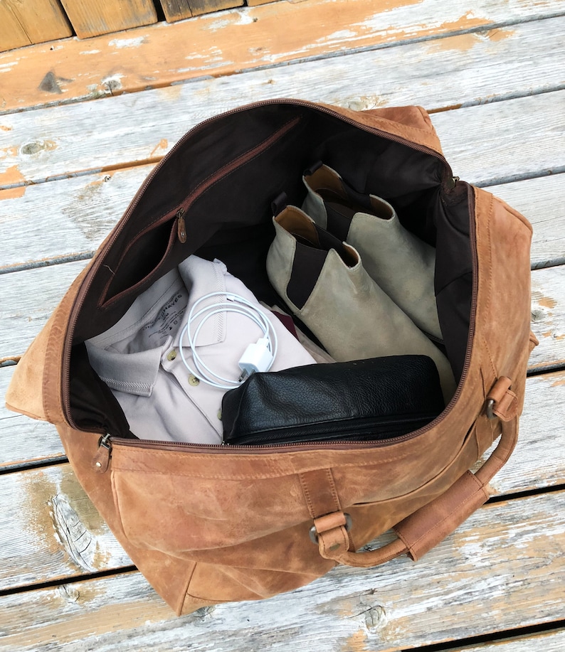 K&ouml;nnte beinhalten: Eine braune Ledertasche mit Rei&szlig;verschluss und einem oberen Griff. Die Tasche ist offen und enth&auml;lt ein wei&szlig;es Hemd, eine schwarze Ledertasche und ein Paar graue Wildlederstiefel.