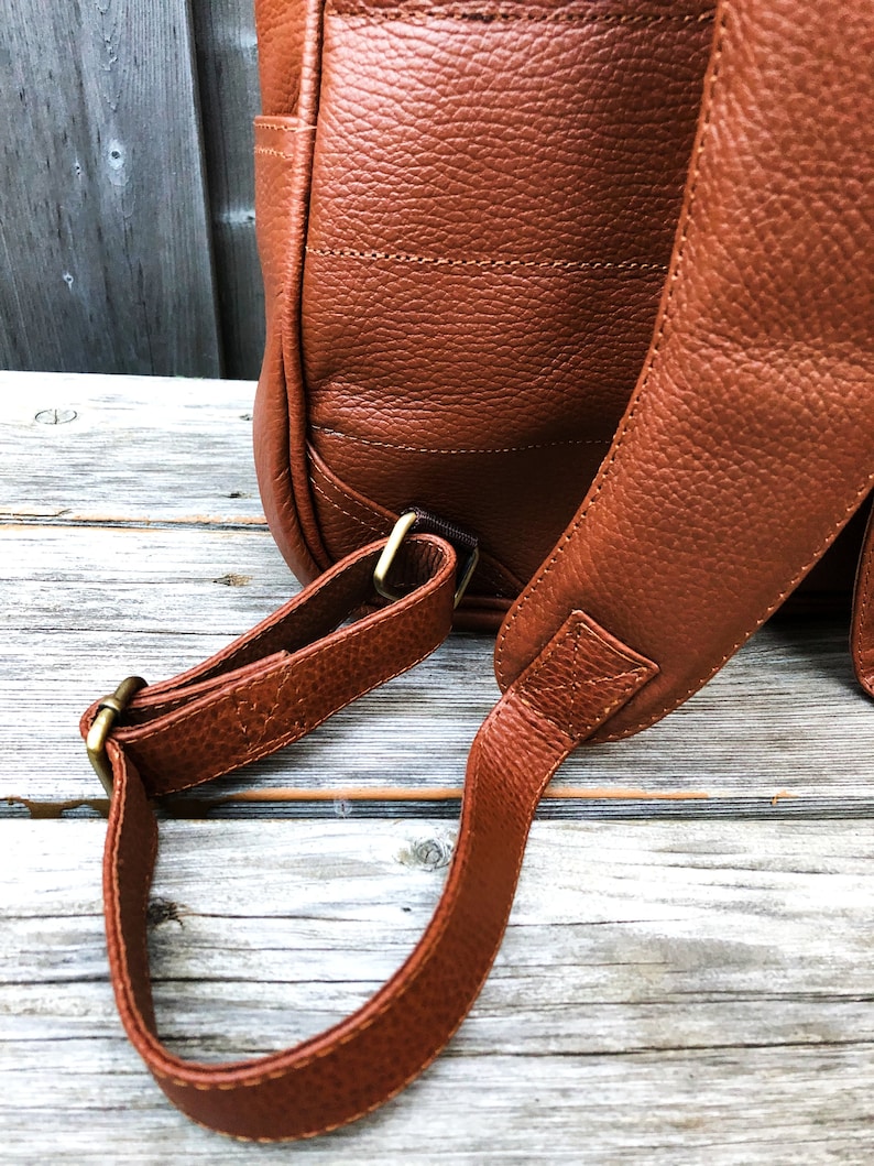 Pu&ograve; includere: Uno zaino in pelle marrone con spalline regolabili e fibbie in metallo dorato. Lo zaino &egrave; realizzato in pelle testurizzata e ha un design classico.