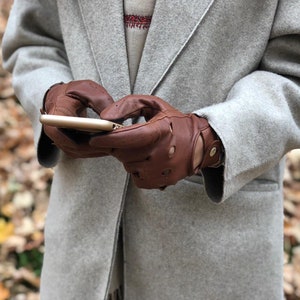 Puede incluir: Una persona con un abrigo gris y guantes de cuero marr&oacute;n est&aacute; usando un tel&eacute;fono inteligente. La persona est&aacute; de pie en un bosque con hojas ca&iacute;das en el suelo.