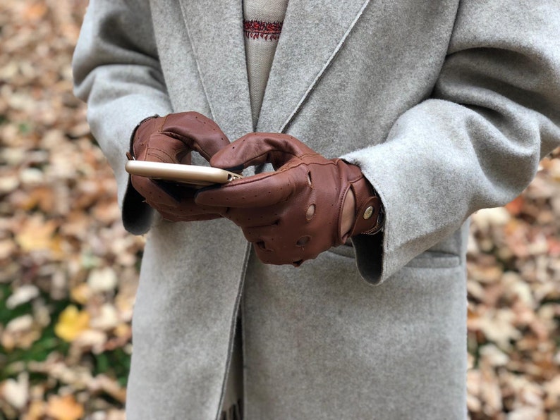 Puede incluir: Una persona con un abrigo gris y guantes de cuero marr&oacute;n est&aacute; usando un tel&eacute;fono inteligente. La persona est&aacute; de pie en un bosque con hojas ca&iacute;das en el suelo.