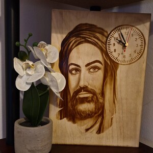 Könnte beinhalten: Eine Holzuhr mit einem Porträt eines Mannes mit Bart und Turban. Das Zifferblatt der Uhr ist weiß mit schwarzen Zahlen und Zeigern. Die Uhr steht auf einem Holzregal.