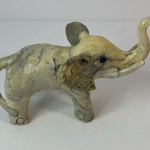 May include: A small, decorative elephant figurine with a raised trunk. The elephant is primarily cream-colored with gray and brown marbling. The ears are a darker shade of gray. The figurine has a detailed, textured appearance, suggesting it is made of a stone-like material.