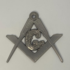 May include: A silver-colored Masonic symbol featuring a square and compass design with ornate detailing. The compass is open, and the square has a ruler along one side. The center features a decorative element. The symbol is likely used for fraternal organizations.