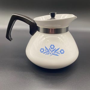 May include: A white ceramic teapot featuring a blue floral pattern on the front. The teapot has a black handle, a black lid knob, and a silver-colored metal band around the neck. The design includes a central blue flower with leaves and stems.