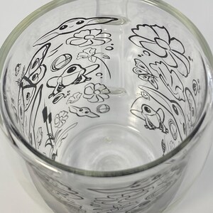 May include: A clear glass jug with a handle, decorated with a black and white pond scene illustration. The design includes frogs, flowers, and butterflies. The jug is empty and rests on a white surface. The jug is made of glass.