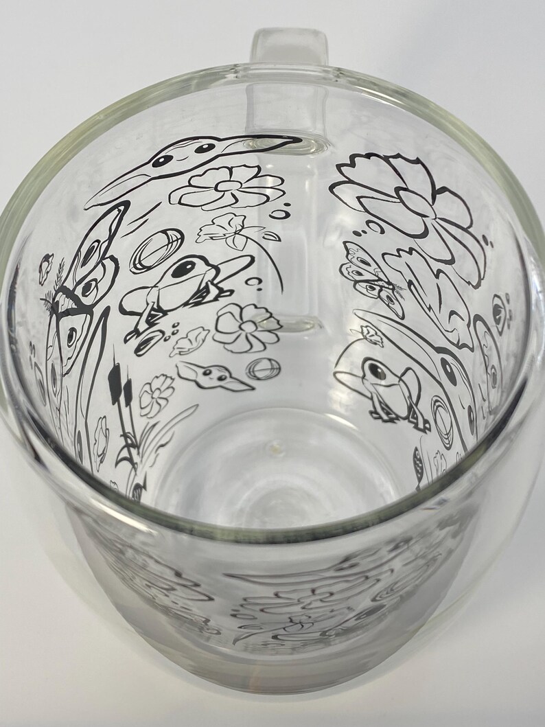 May include: A clear glass jug with a handle, decorated with a black and white pond scene illustration. The design includes frogs, flowers, and butterflies. The jug is empty and rests on a white surface. The jug is made of glass.