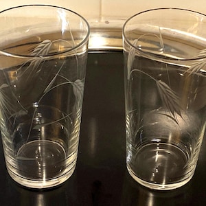 May include: Two clear, etched glass tumblers with a wheat stalk design. The glasses are cylindrical with a slightly flared rim. The etched design is delicate and adds a touch of elegance to the glassware. The glasses are empty and sit on a dark surface.