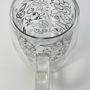 May include: A clear glass mug with a double-walled design. The interior features a whimsical black and white illustration of flowers, butterflies, and birds. The mug has a clear handle and is suitable for hot or cold drinks.