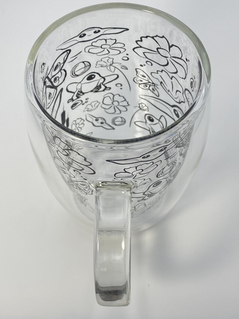 May include: A clear glass mug with a double-walled design. The interior features a whimsical black and white illustration of flowers, butterflies, and birds. The mug has a clear handle and is suitable for hot or cold drinks.