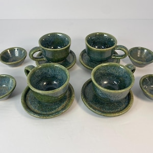 May include: A set of handmade ceramic teacups, saucers, and small bowls. The set includes four teacups with handles, four saucers, and four small bowls. The ceramic pieces have a speckled blue and green glaze.