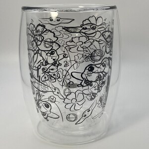 May include: A clear double-walled glass featuring a black line art design of flowers and a cartoon character. The glass has a rounded base and a slightly flared rim. The design is intricate and covers the entire surface.