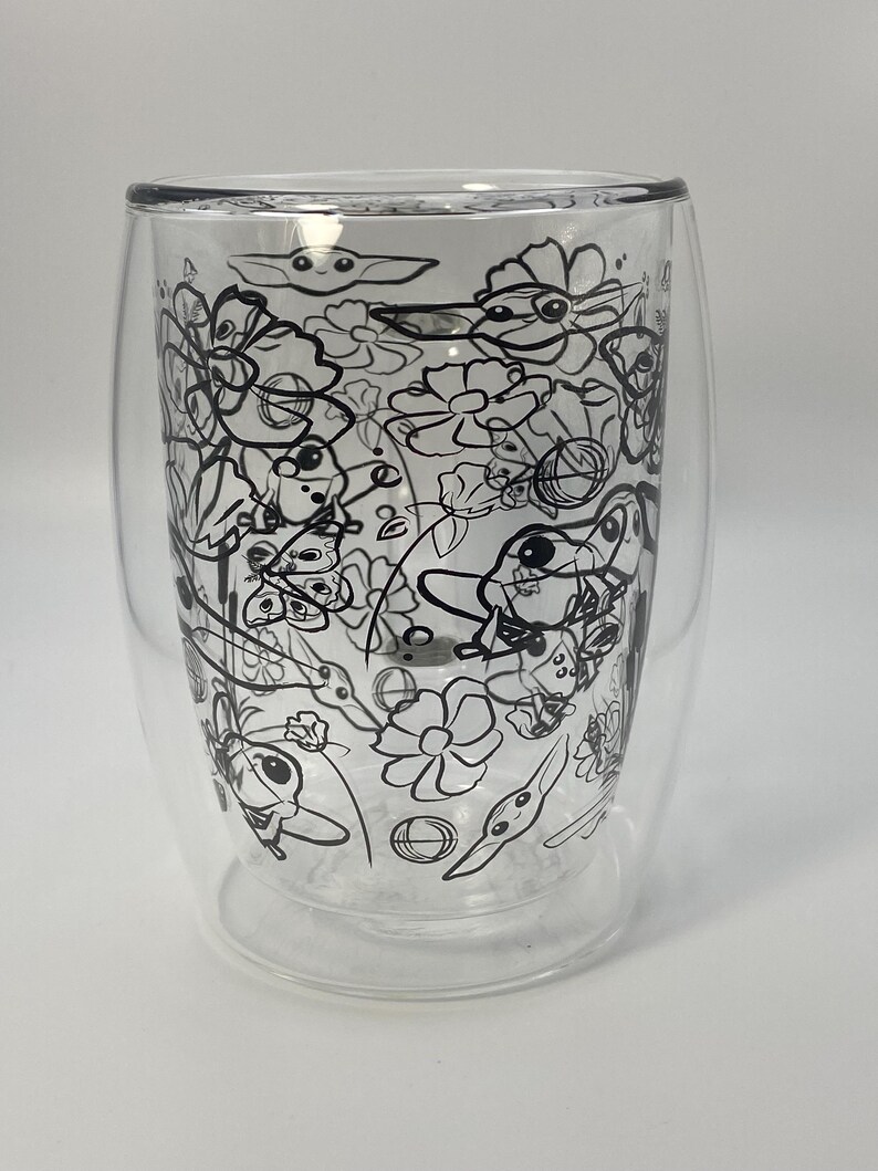 May include: A clear double-walled glass featuring a black line art design of flowers and a cartoon character. The glass has a rounded base and a slightly flared rim. The design is intricate and covers the entire surface.