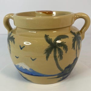 May include: Tan ceramic jar with two handles and a lid. The jar is decorated with a painted tropical scene featuring palm trees, blue waves, and a white mountain. The lid has a small brown knob. The jar is likely used for storage.