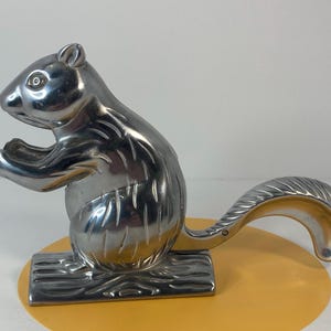 May include: A silver-colored squirrel nutcracker. The nutcracker is in the shape of a squirrel, with its tail forming the handle. The squirrel is standing on a rectangular base that resembles a log.