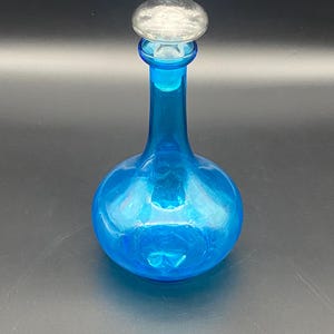 May include: A vibrant blue glass decanter with a rounded base and a slender neck. The vessel is topped with a clear, circular glass stopper that fits securely into the opening. The glass is translucent, allowing light to pass through the blue material.