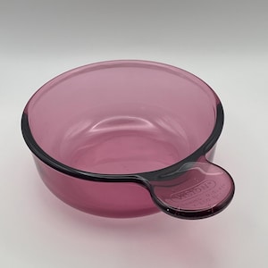May include: A clear, round, pink glass bowl with a handle. The bowl has a black rim and handle. The handle has the word "VISION" and other text printed on it. The bowl is sitting on a white surface.