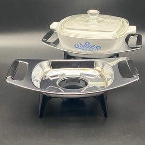 May include: A white rectangular casserole dish with a clear glass lid and blue floral pattern, resting on a chrome metal stand with black handles. In the foreground, a second empty chrome metal stand with black handles and black legs.
