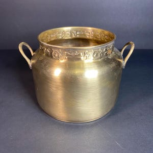 May include: A gold-colored metal bowl with two small handles on the sides. The rim of the bowl has a decorative pattern. The bowl has a rounded shape and sits on a dark surface.