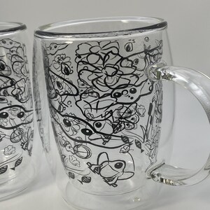 May include: Two clear glass mugs with a double-walled design. Each mug has a black and white doodle pattern of flowers, fish, and other whimsical shapes. The mugs have clear handles and are set against a white background.
