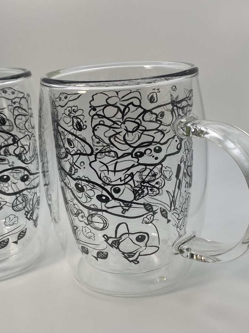 May include: Two clear glass mugs with a double-walled design. Each mug has a black and white doodle pattern of flowers, fish, and other whimsical shapes. The mugs have clear handles and are set against a white background.