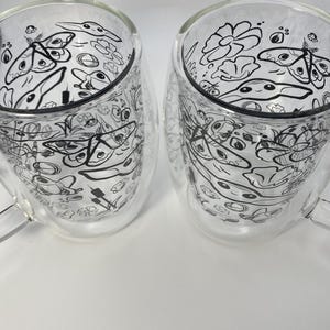 May include: Two clear glass mugs with a double-walled design. Each mug has a black and white doodle pattern of various shapes and creatures. The mugs have a handle and are set against a white background.