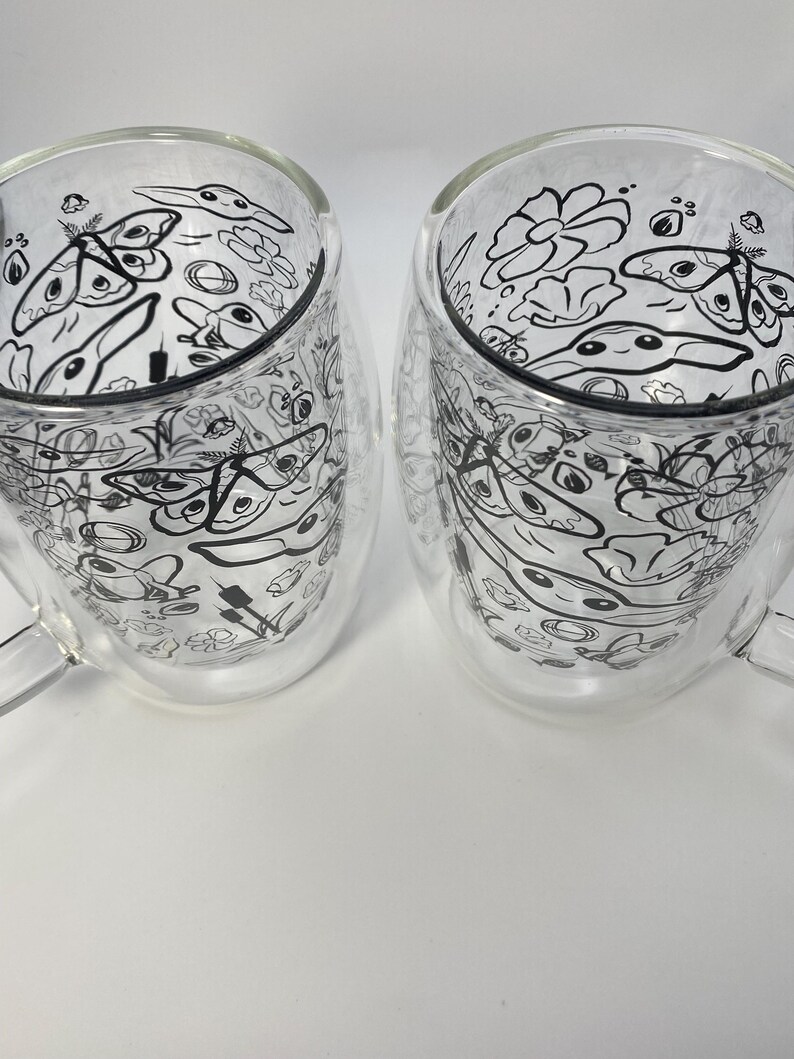 May include: Two clear glass mugs with a double-walled design. Each mug has a black and white doodle pattern of various shapes and creatures. The mugs have a handle and are set against a white background.