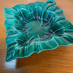 May include: A ceramic leaf-shaped dish with a glossy, emerald green glaze. The dish features detailed veining and a small handle. The center of the dish has a darker, shaded area, adding depth to the design. A decorative piece for serving or display.