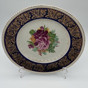May include: A round ceramic plate featuring a central bouquet of purple, pink, and yellow roses with green leaves. The plate has a wide, deep blue border decorated with intricate gold filigree patterns and a scalloped, gold-trimmed edge.