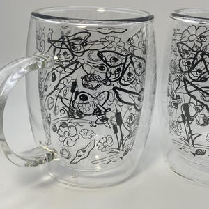 May include: Two clear glass mugs with a double-walled design. Each mug has a black and white floral and nature-themed pattern. The mugs have a handle and are suitable for hot or cold drinks.