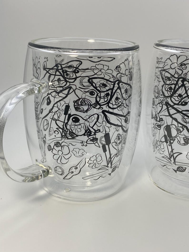 May include: Two clear glass mugs with a double-walled design. Each mug has a black and white floral and nature-themed pattern. The mugs have a handle and are suitable for hot or cold drinks.