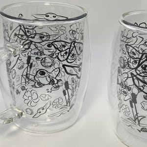 May include: Two clear glass mugs with a double-walled design. Each mug has a black and white doodle pattern of a character, flowers, and other shapes. One mug has a handle. The mugs are set against a white backdrop.
