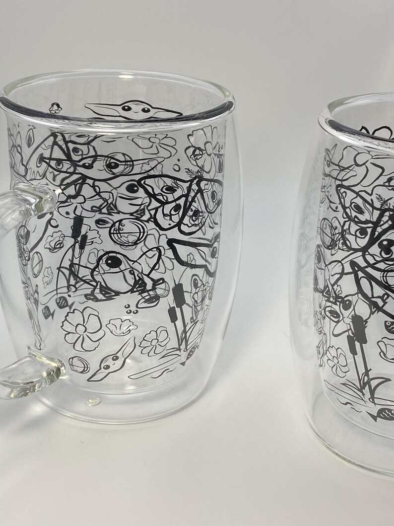 May include: Two clear glass mugs with a double-walled design. Each mug has a black and white doodle pattern of a character, flowers, and other shapes. One mug has a handle. The mugs are set against a white backdrop.