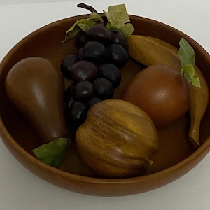 May include: A wooden bowl filled with decorative wooden fruit. The bowl contains a bunch of dark purple grapes, a pear, a peach, a banana, and a uniquely shaped fruit. The bowl and fruit are various shades of brown.
