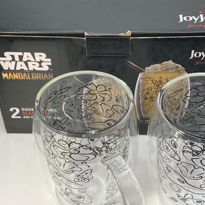 May include: Two double-walled glass mugs with handles, featuring a black and white Star Wars Mandalorian design. Each mug holds 400 ml. The mugs are displayed in front of a black box with the Star Wars Mandalorian logo.
